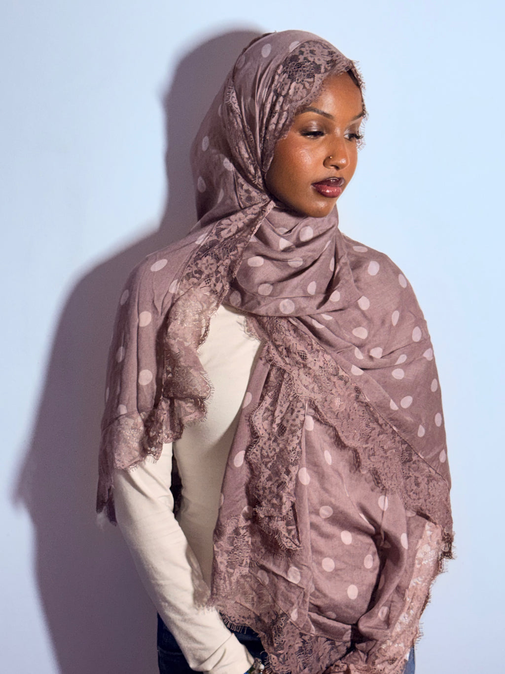 A woman wearing a dusty mauve hijab with white polka dots and lace trim, styled over a cream long-sleeve top against a light blue background.